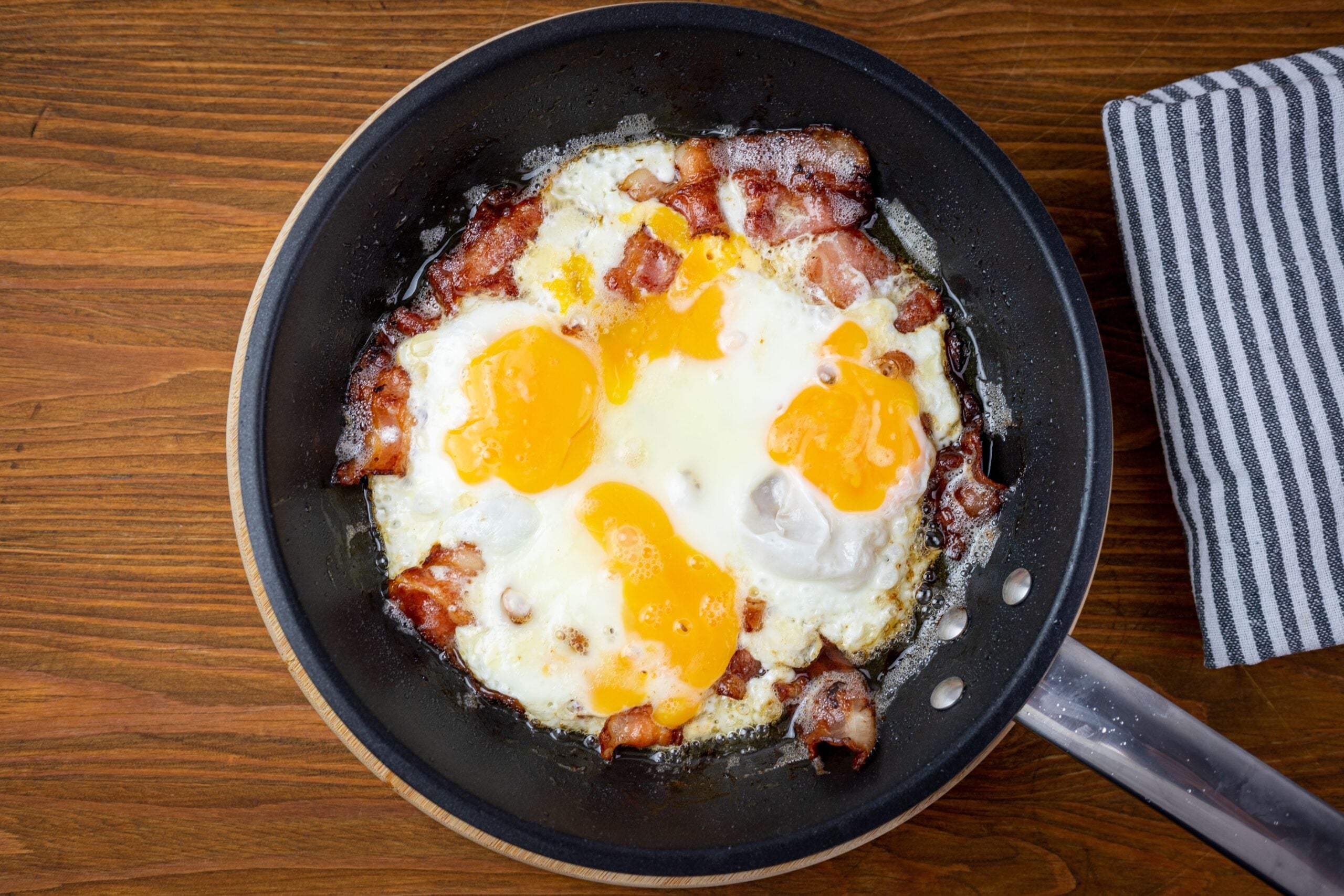 Three sunny-side-up eggs and pieces of bacon cooking in a black frying pan on a wooden surface, with a striped cloth to the side.
