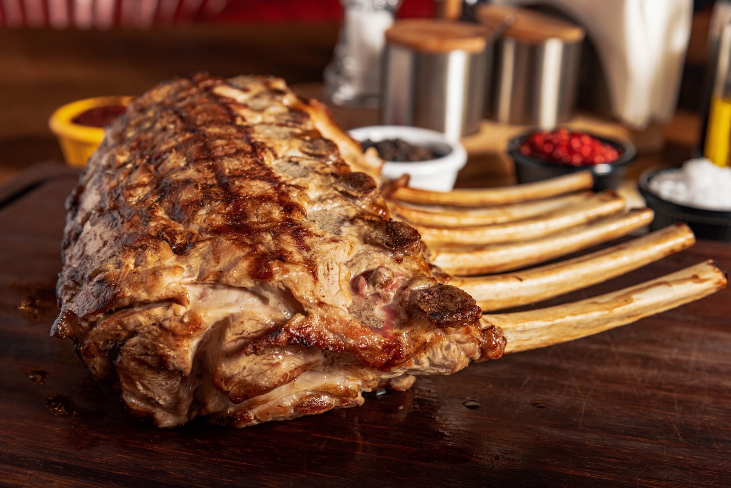 A grilled rack of ribs rests on a wooden board, with seasonings and condiments in small bowls blurred in the background.