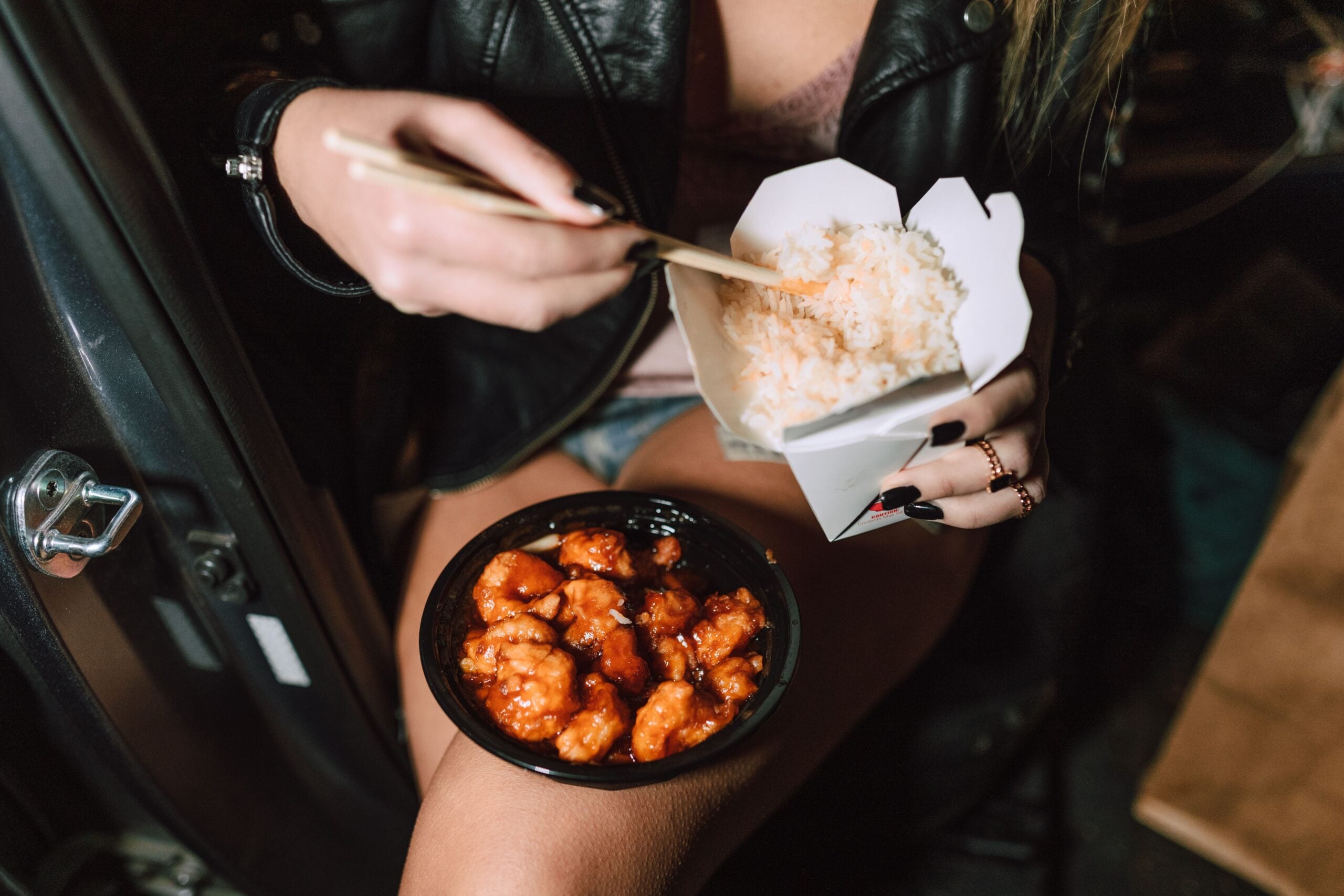 A person sitting with a takeout container of white rice and a black bowl of saucy chicken, eating with chopsticks while wearing a leather jacket and shorts.