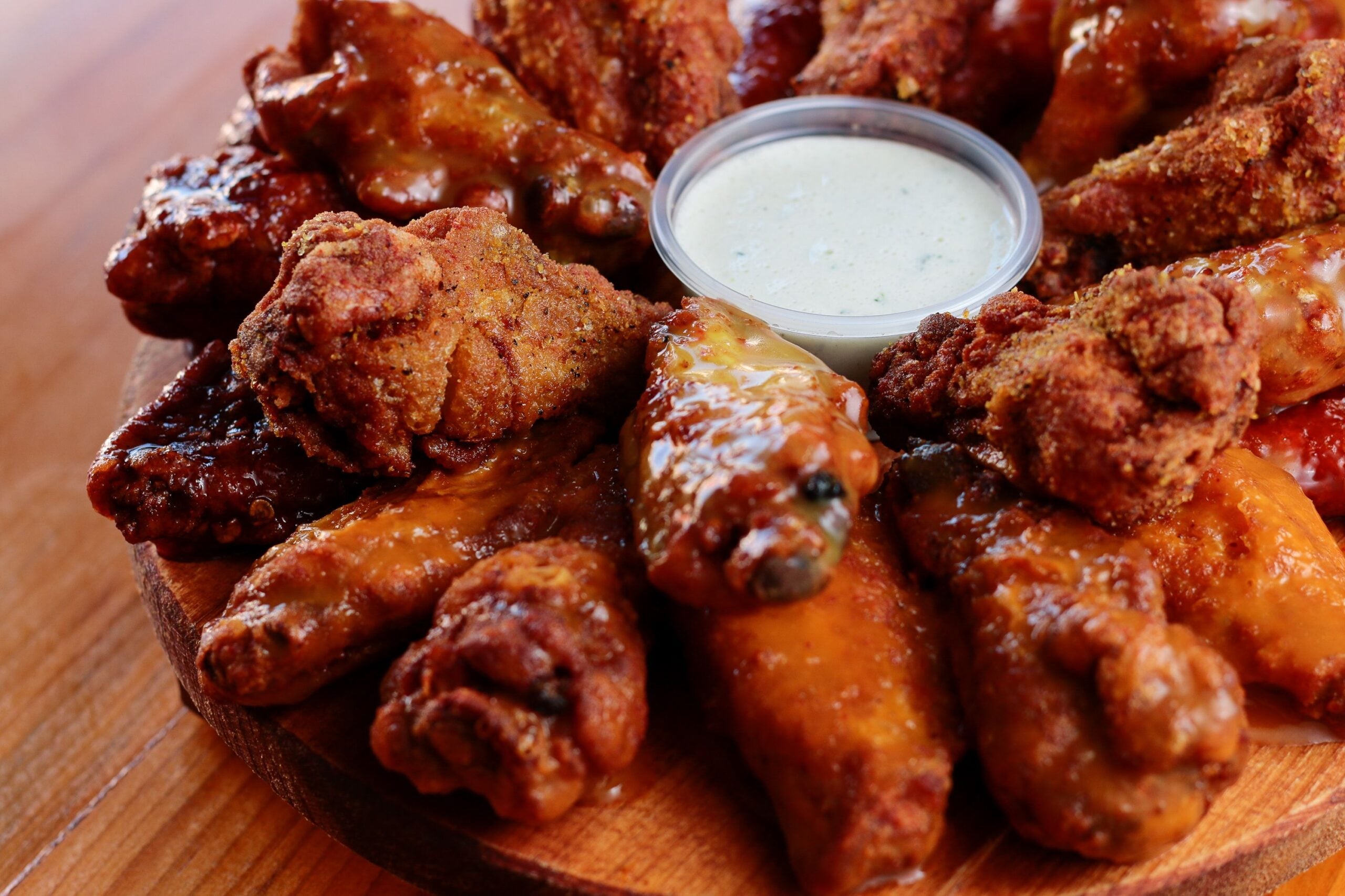 A round wooden platter filled with various sauced and seasoned chicken wings arranged around a cup of creamy dipping sauce, placed on a wooden table.