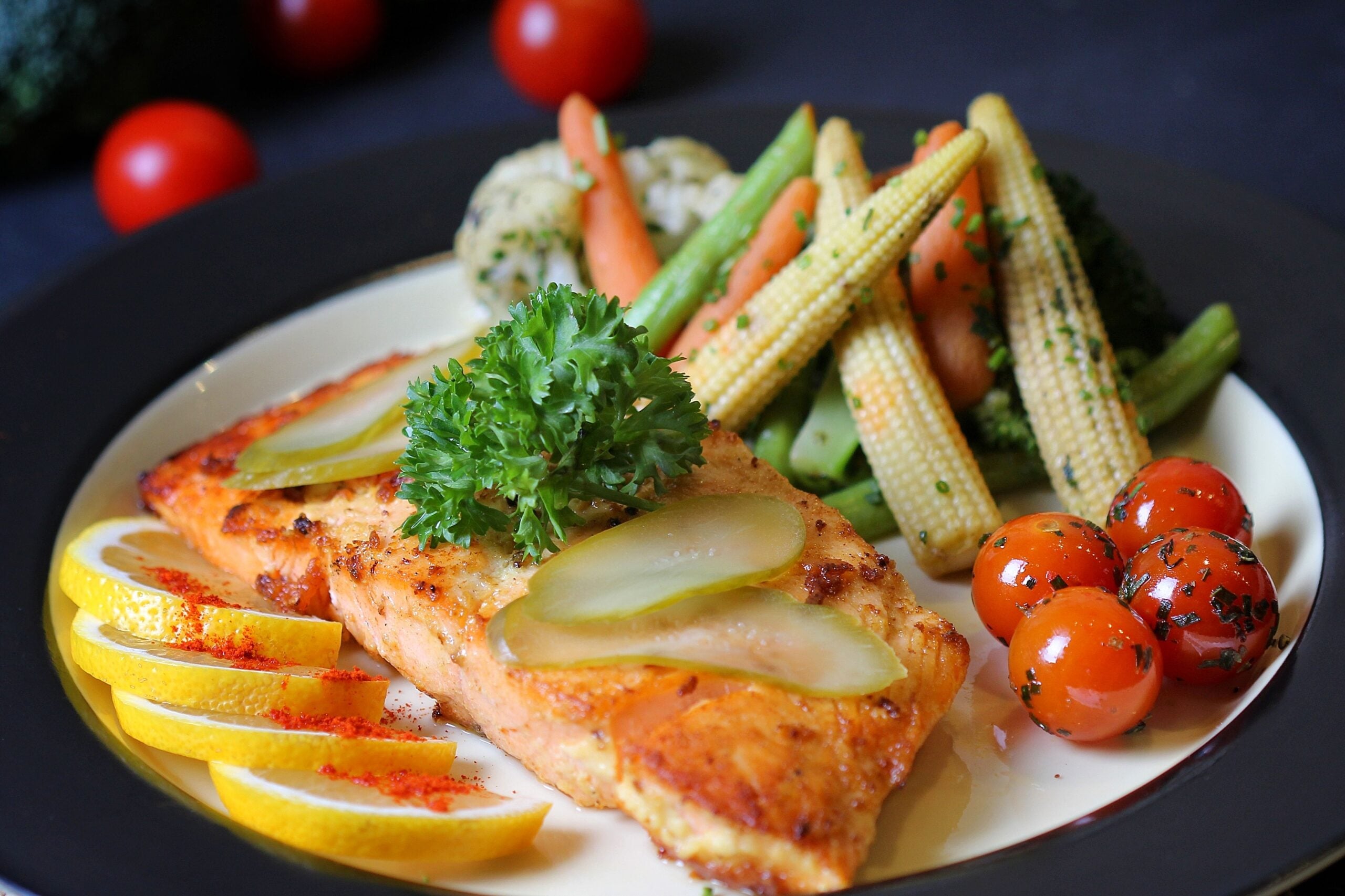 Grilled salmon fillet garnished with parsley and pickles, served with lemon wedges, cherry tomatoes, baby corn, carrots, broccoli, and rice.