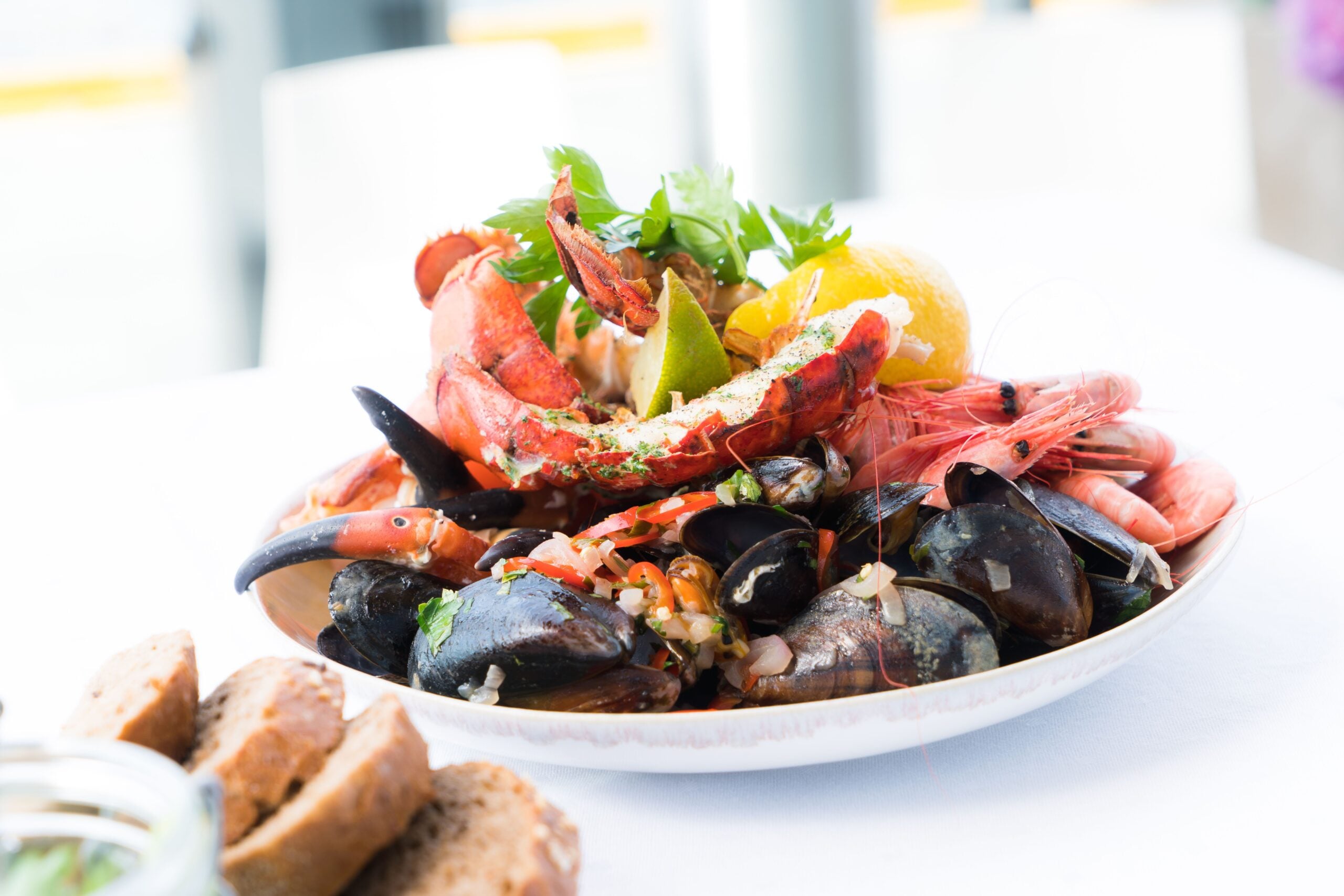 A plate of assorted seafood including lobster, shrimp, mussels, and clams, garnished with herbs and a lemon wedge, sits on a white table next to slices of brown bread.