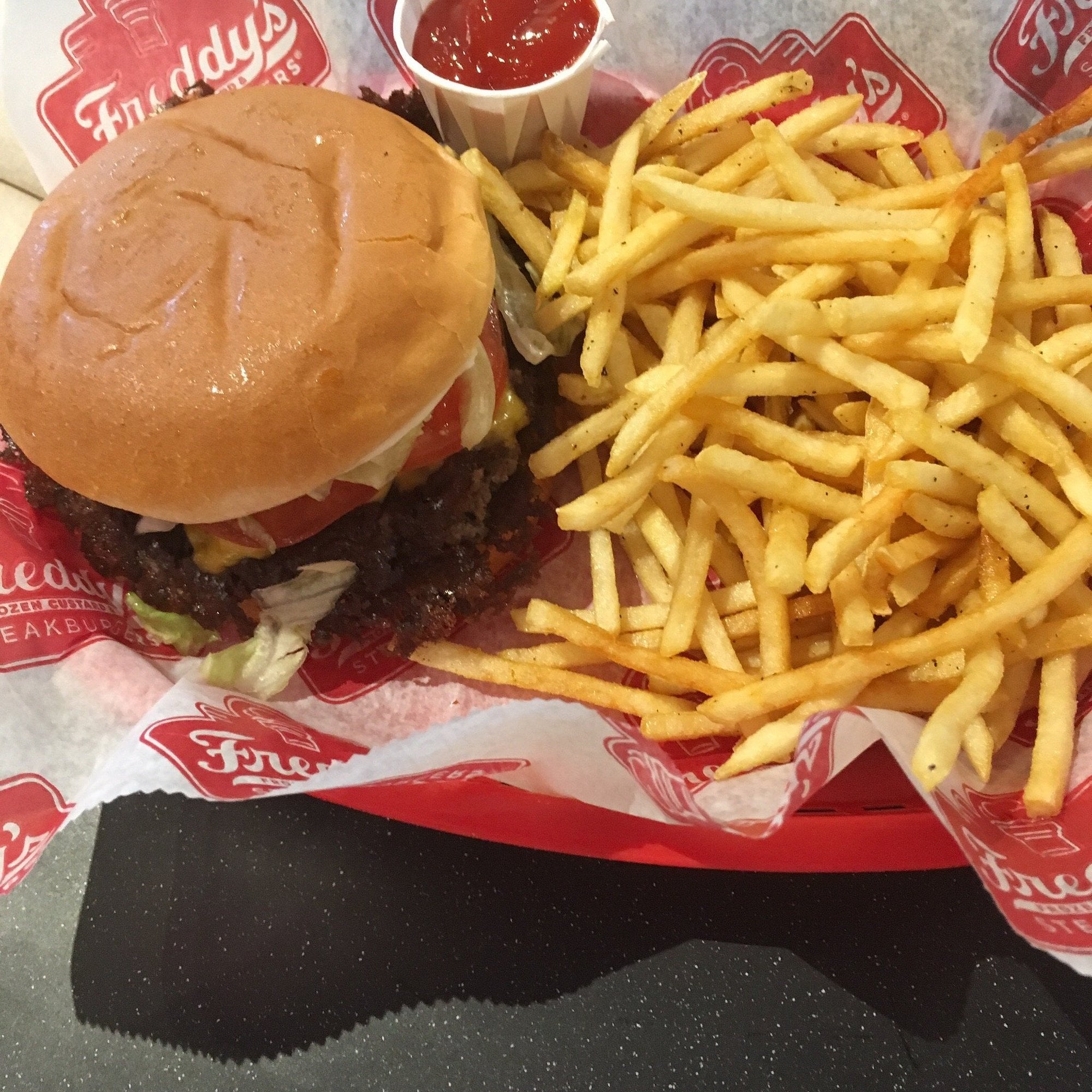 A cheeseburger with lettuce, tomato, and sauce sits next to a large serving of thin French fries and ketchup in a red tray lined with Freddy’s branded paper, showcasing the classic appeal of regional fast-food chains.