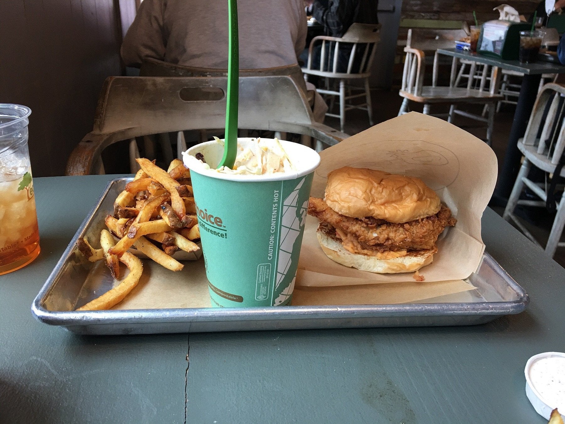A tray with seasoned fries, a fried chicken sandwich on a bun, and a cup of milkshake with a green straw on a table in a rustic-style restaurant. An iced drink is beside the tray.
