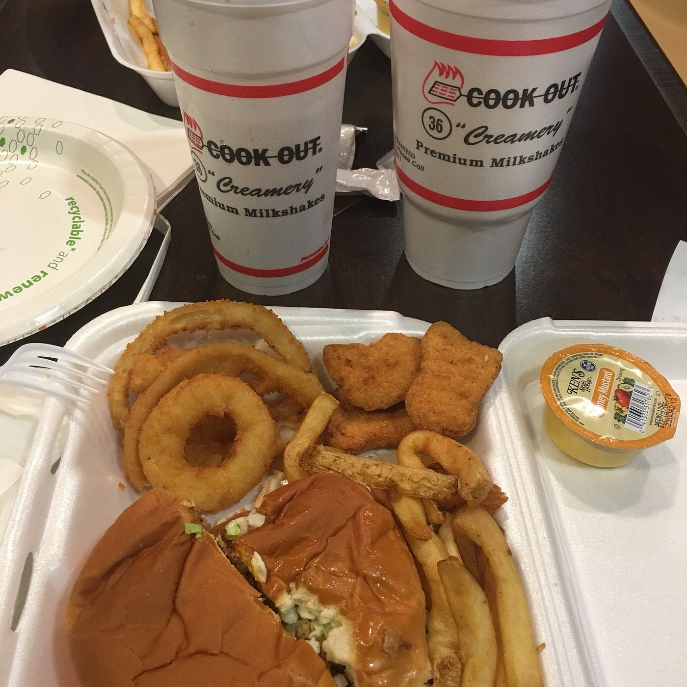 A takeout meal from one of the top regional fast-food chains, featuring a burger, fries, onion rings, nuggets, and sauce in a foam container, plus two large Cook Out milkshake cups on a table.