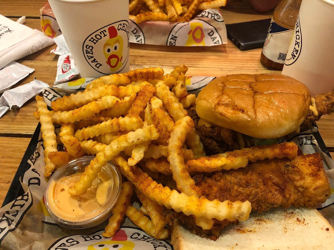 A tray with crinkle-cut fries, dipping sauce, a fried chicken sandwich, and chicken tenders—classic fare from one of the most popular regional fast-food chains. Two drinks and more fries are in the background. The tray paper says “Dave’s Hot Chicken.”