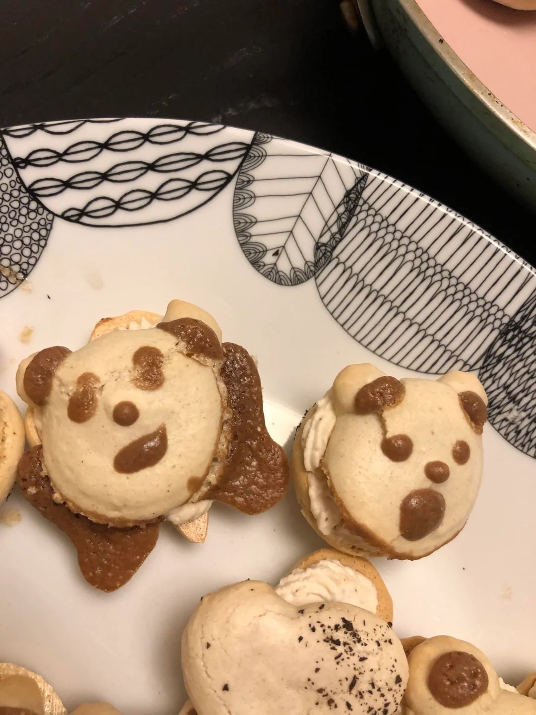 Two macarons decorated to look like bear faces with chocolate features are on a patterned white plate. One bear has a smiling face and the other has a neutral expression. Another macaron is partially visible below them.