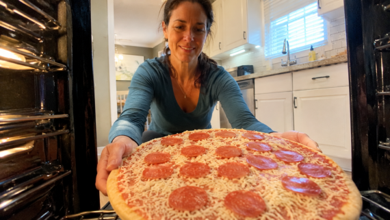A woman is placing a pepperoni pizza from one of her favorite frozen pizza brands into an oven, viewed from inside the oven, in a bright kitchen with white cabinets and countertops.