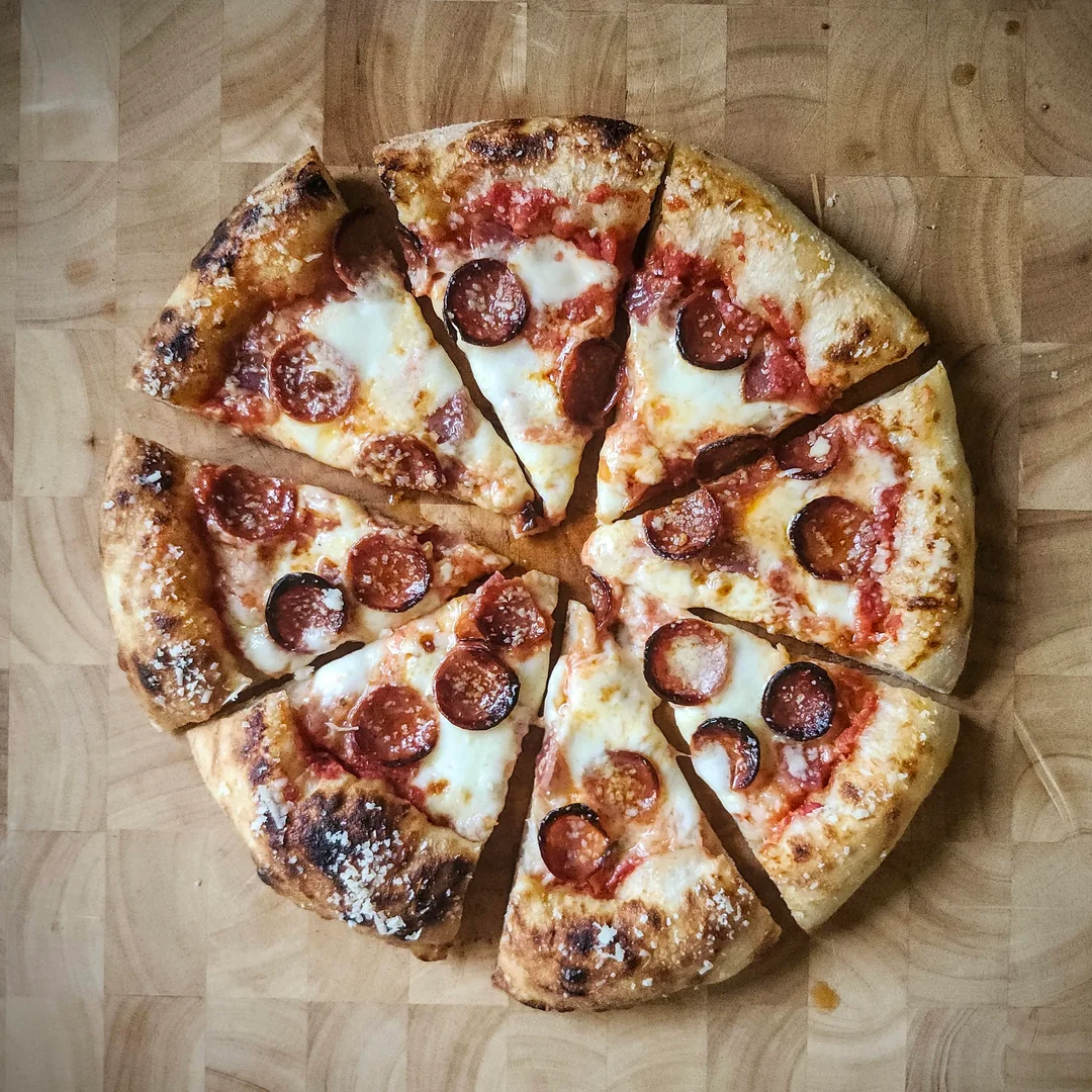 A pepperoni pizza cut into eight slices sits on a wooden surface. The crust is golden and slightly charred, with melted cheese and pepperoni slices scattered on top.