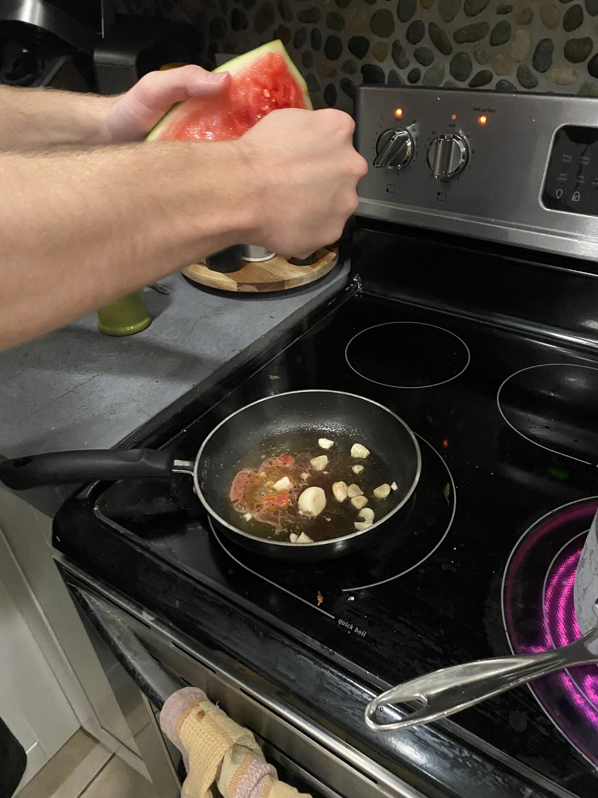 A person holds a watermelon slice over a frying pan with oil, garlic, and herbs, cooking on a stove with a pot nearby.