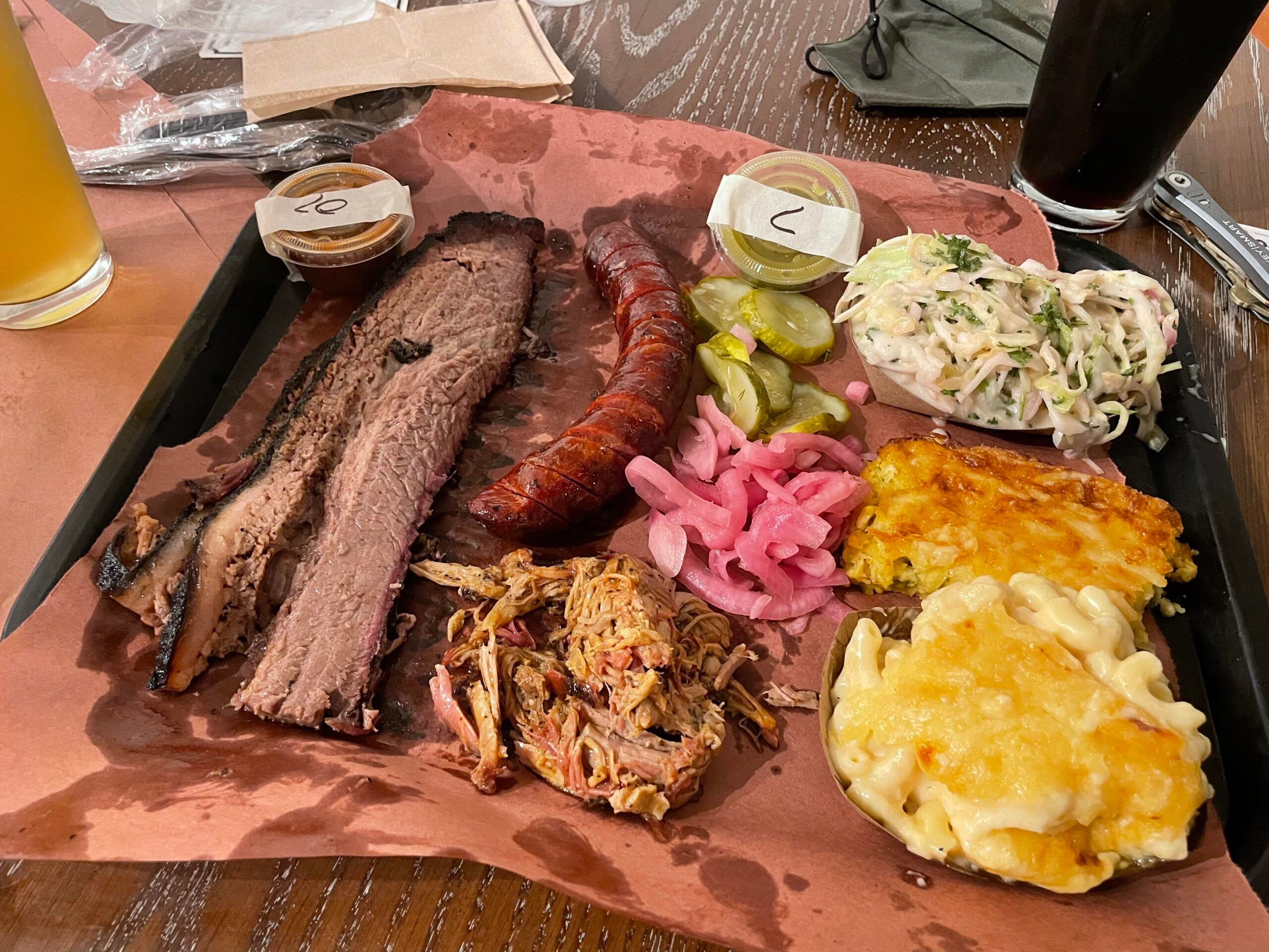 A tray of barbecue food with brisket, sausage, pulled pork, pickles, pickled onions, coleslaw, macaroni and cheese, cheesy potatoes, and sauces, served on brown paper with drinks nearby.