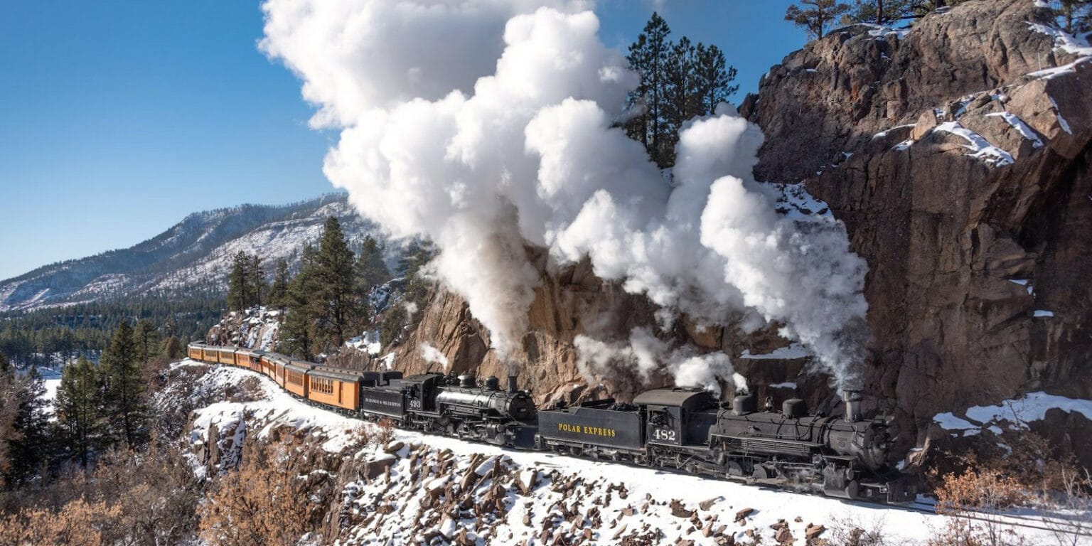 A black steam locomotive labeled “Polar Express” pulls yellow passenger cars through snowy mountains, with thick white smoke billowing behind it against a clear sky—capturing the enchanting spirit found in the best Christmas towns in America.