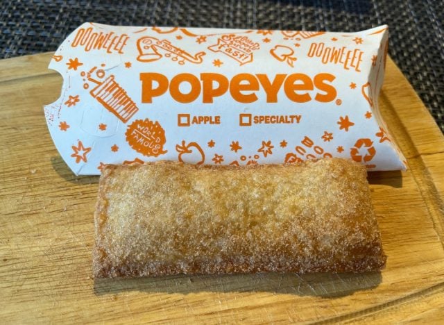 A Popeyes apple pie in a crispy, golden-brown crust sits on a wooden surface, with its branded orange and white packaging labeled “APPLE” and “POPEYES” behind it.