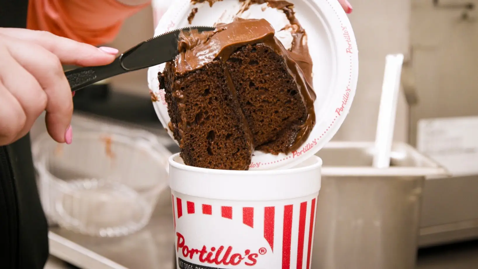 A hand uses a knife to drop a slice of chocolate cake with frosting from a plate into a large Portillo's cup with red stripes in a kitchen setting.