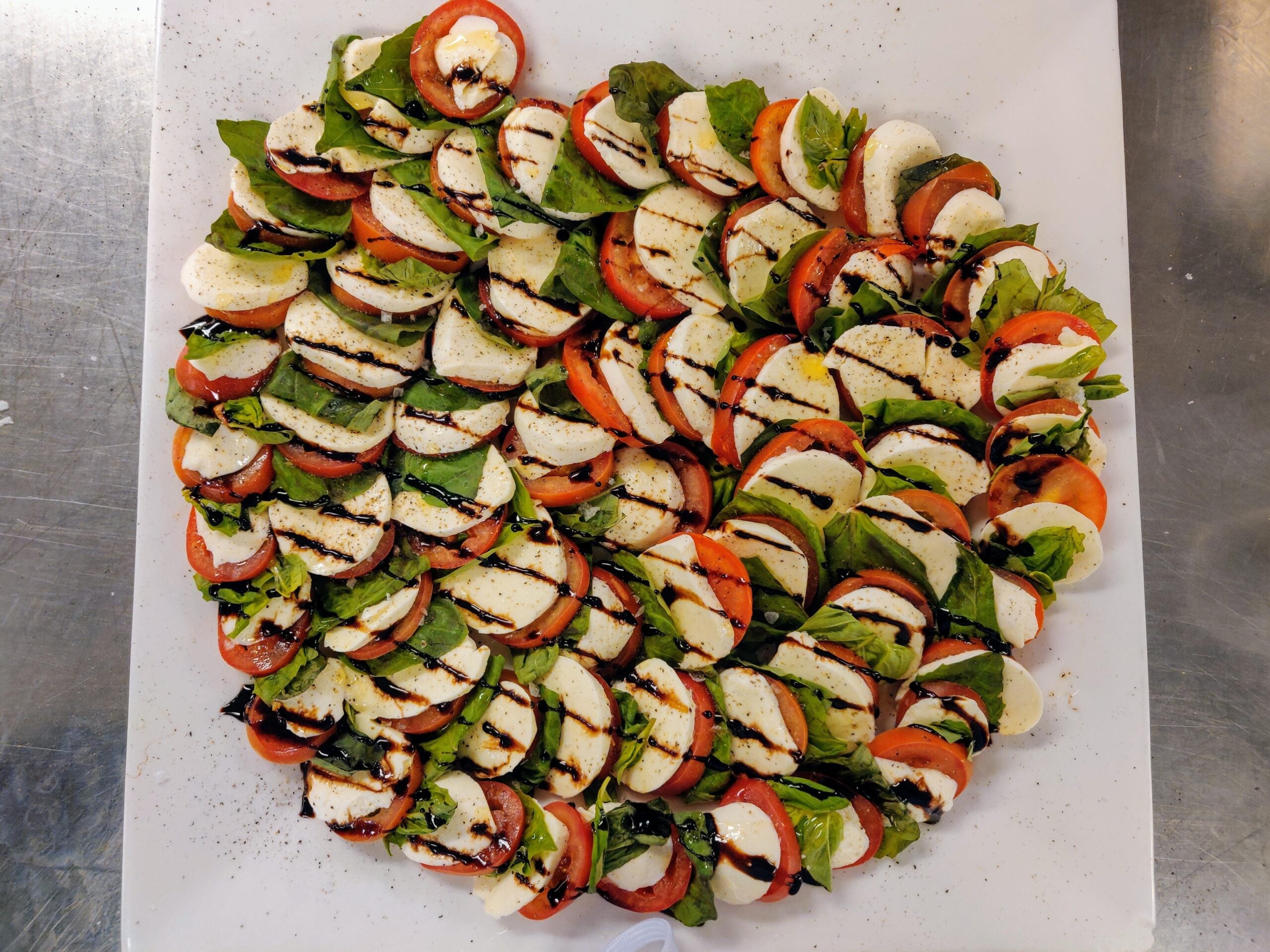 Rows of sliced tomatoes, fresh mozzarella, and basil leaves arranged in a heart shape, drizzled with balsamic glaze, on a white surface.