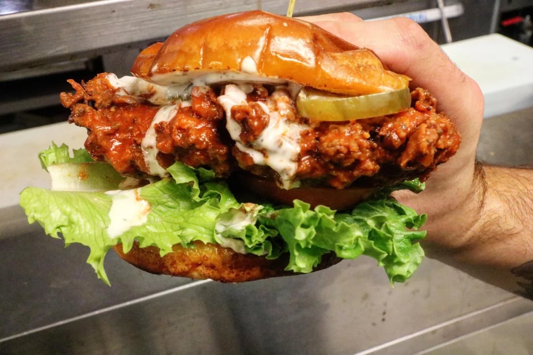 A hand holds a large fried chicken sandwich with lettuce, pickles, and creamy sauce on a toasted bun.