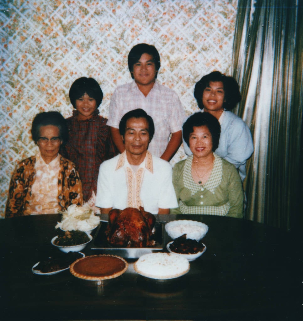 A vintage family portrait featuring six people gathered around a table spread with a festive meal, including a roasted chicken, pies, and bowls of food—a timeless snapshot of nostalgic dinners. They sit in front of floral wallpaper and curtains, all smiling at the camera.
