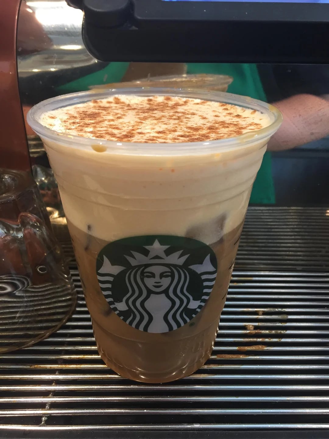 A plastic cup of iced coffee, reminiscent of fast-food grocery store products, sits on a metal counter topped with foam and cinnamon. The cup has a Starbucks logo on the front.