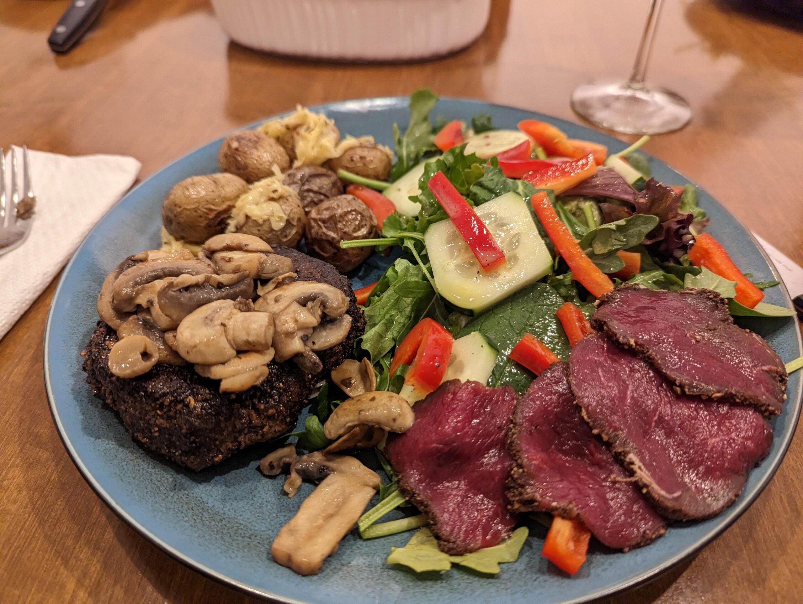 A plate with sliced cured meat on greens, a mound of sautéed mushrooms on a dark patty, roasted potatoes, and a salad with cucumber and red bell pepper. A fork, napkin, and wine glass are nearby.