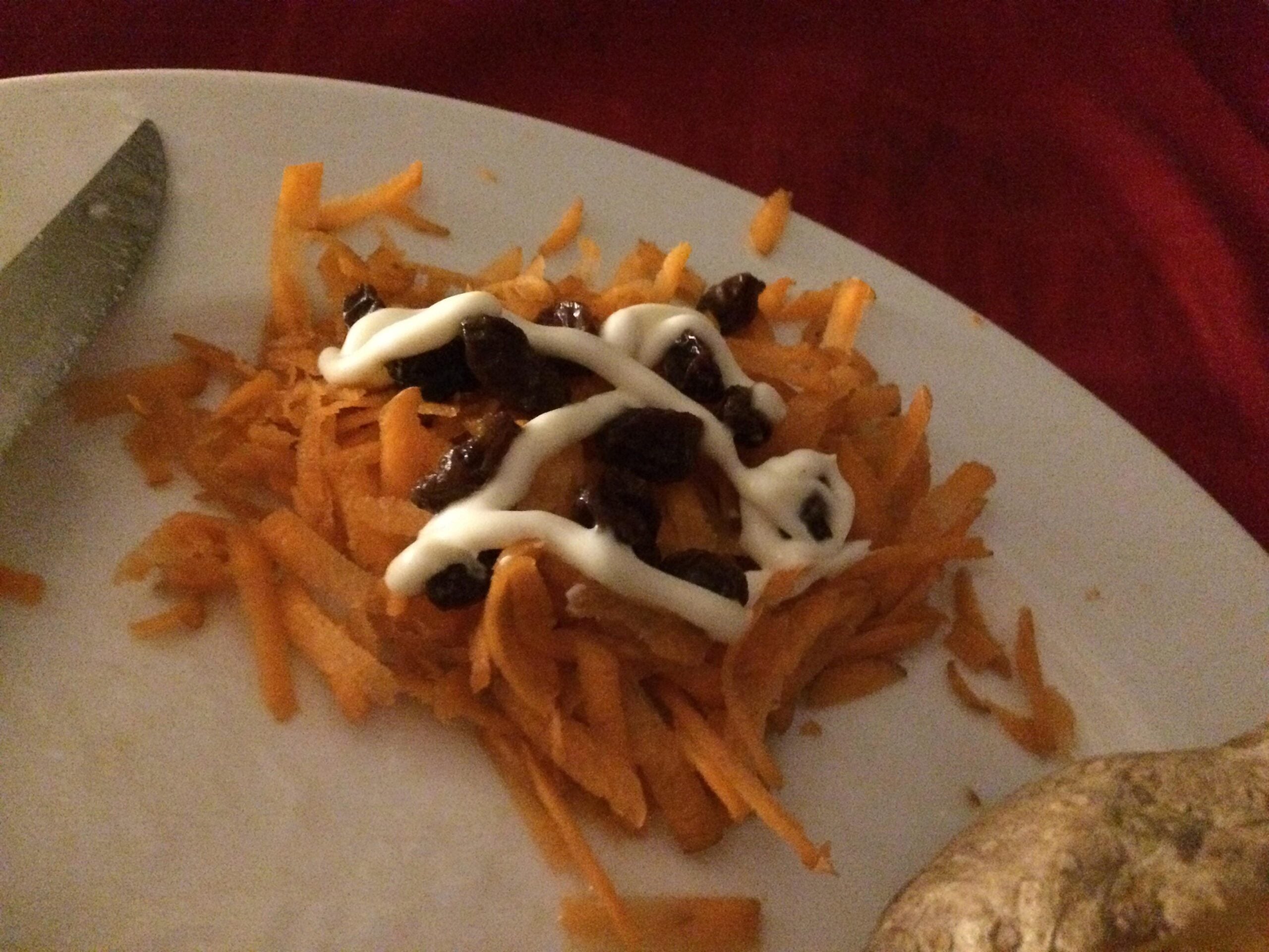 A pile of shredded carrots topped with raisins and a drizzle of mayonnaise sits on a white plate, next to a knife and part of a baked potato.