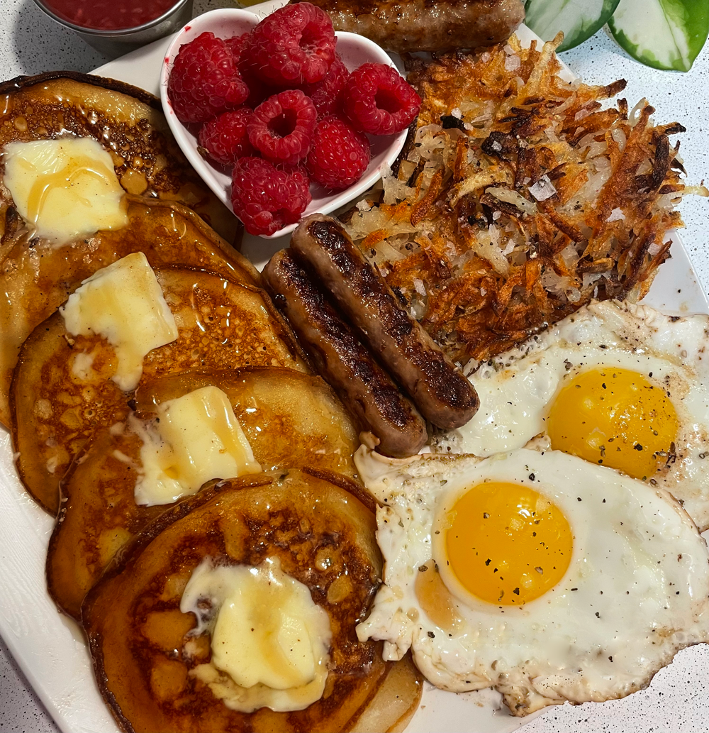 A breakfast plate featuring fluffy homemade pancakes topped with butter, sunny-side-up eggs, hash browns, sausages, fresh raspberries, and a small side of sauce.