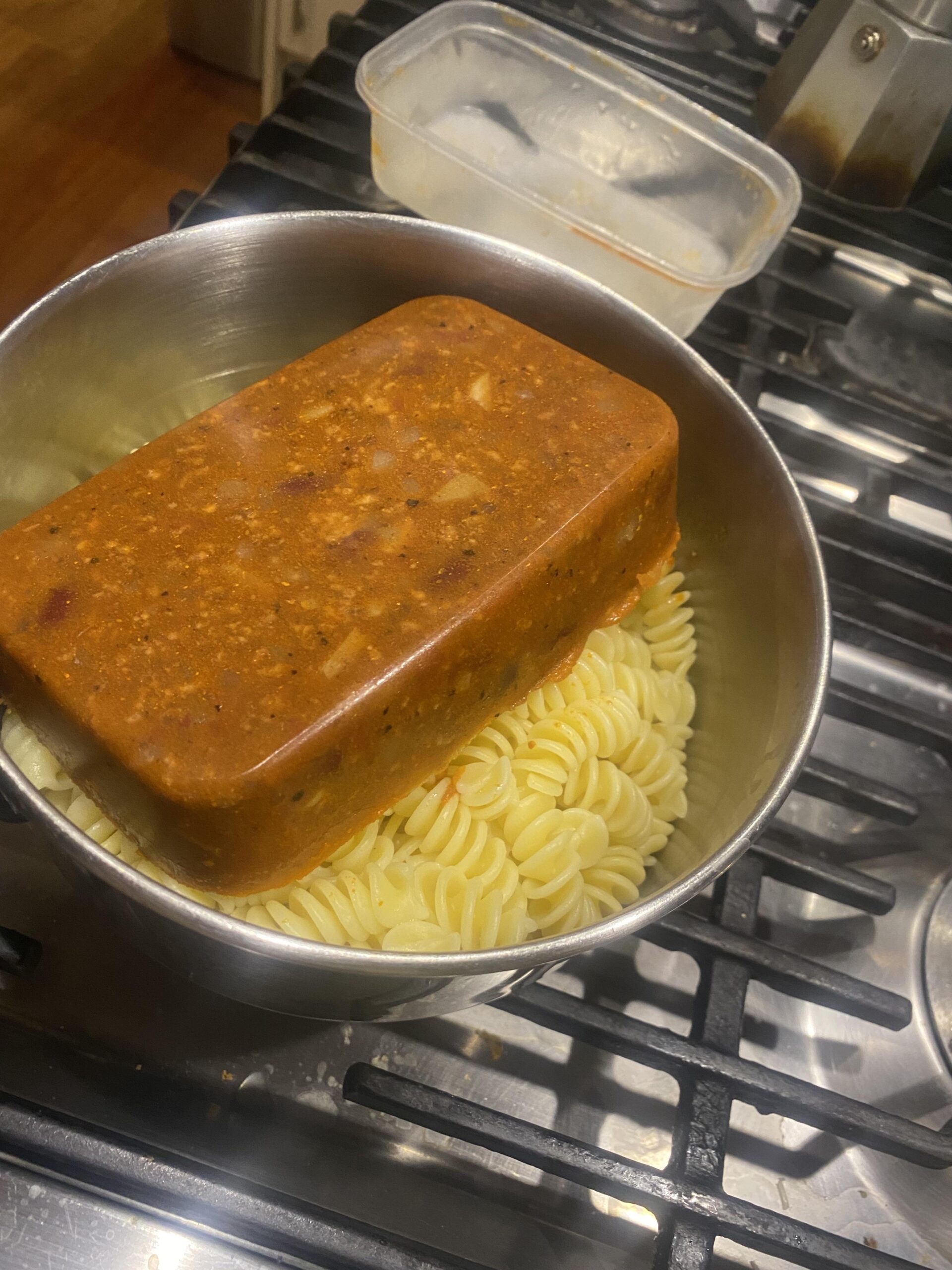 A solid block of frozen sauce sits on top of cooked rotini pasta in a pot on a stovetop. An empty plastic container is nearby.