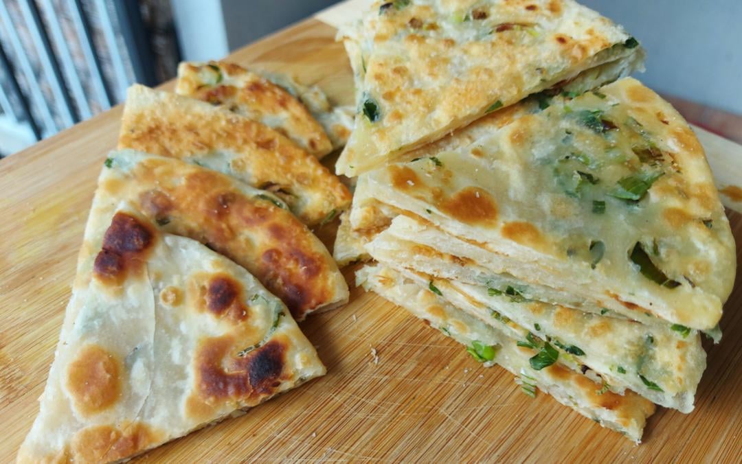 Golden, crispy Chinese homemade pancakes cut into wedges, stacked neatly on a wooden cutting board. The pancakes show layers with green scallions visible inside.