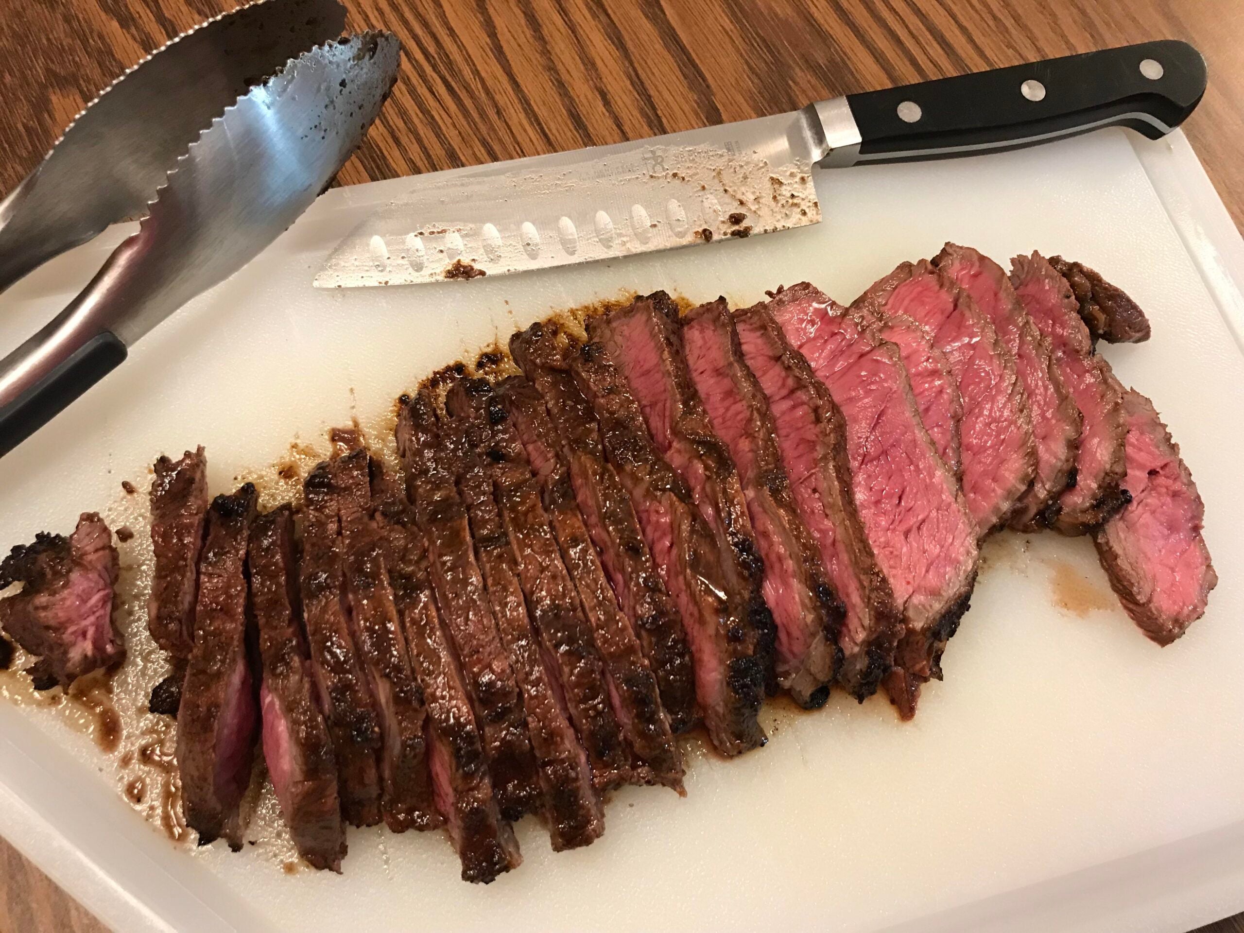 Sliced medium-rare steak on a white cutting board with a knife and metal tongs beside it, showing juicy pink centers and browned edges.