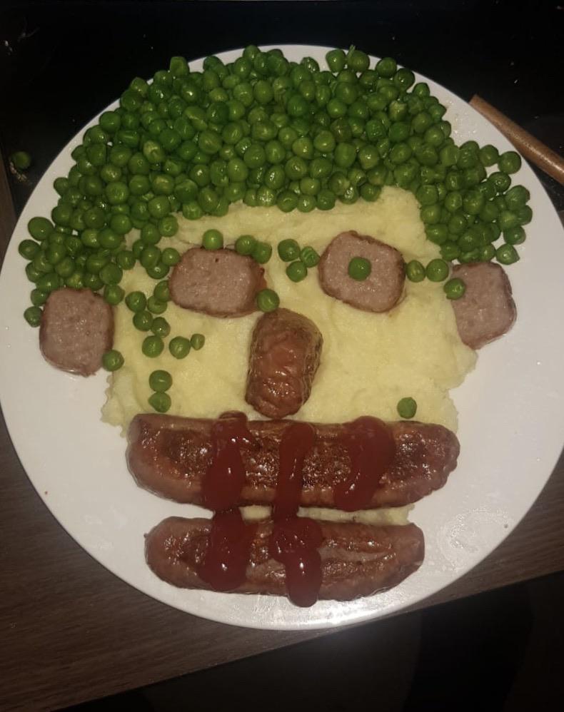 A plate arranged to look like a face, with mashed potatoes as the face, peas as hair, sausages as the mouth with ketchup, meat pieces as eyes and nose, and peas for pupils—perfect for those moments when hilarious cooking disasters turn into creative masterpieces.