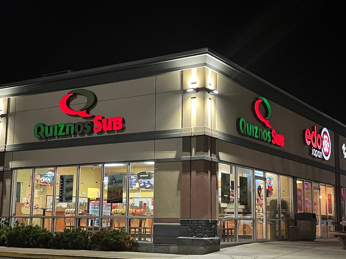 Fast-food chains from the '90s. A brightly lit Quiznos Sub and Edo Japan restaurant at night, with illuminated signs, large windows, and visible seating inside. The parking lot in front is empty.