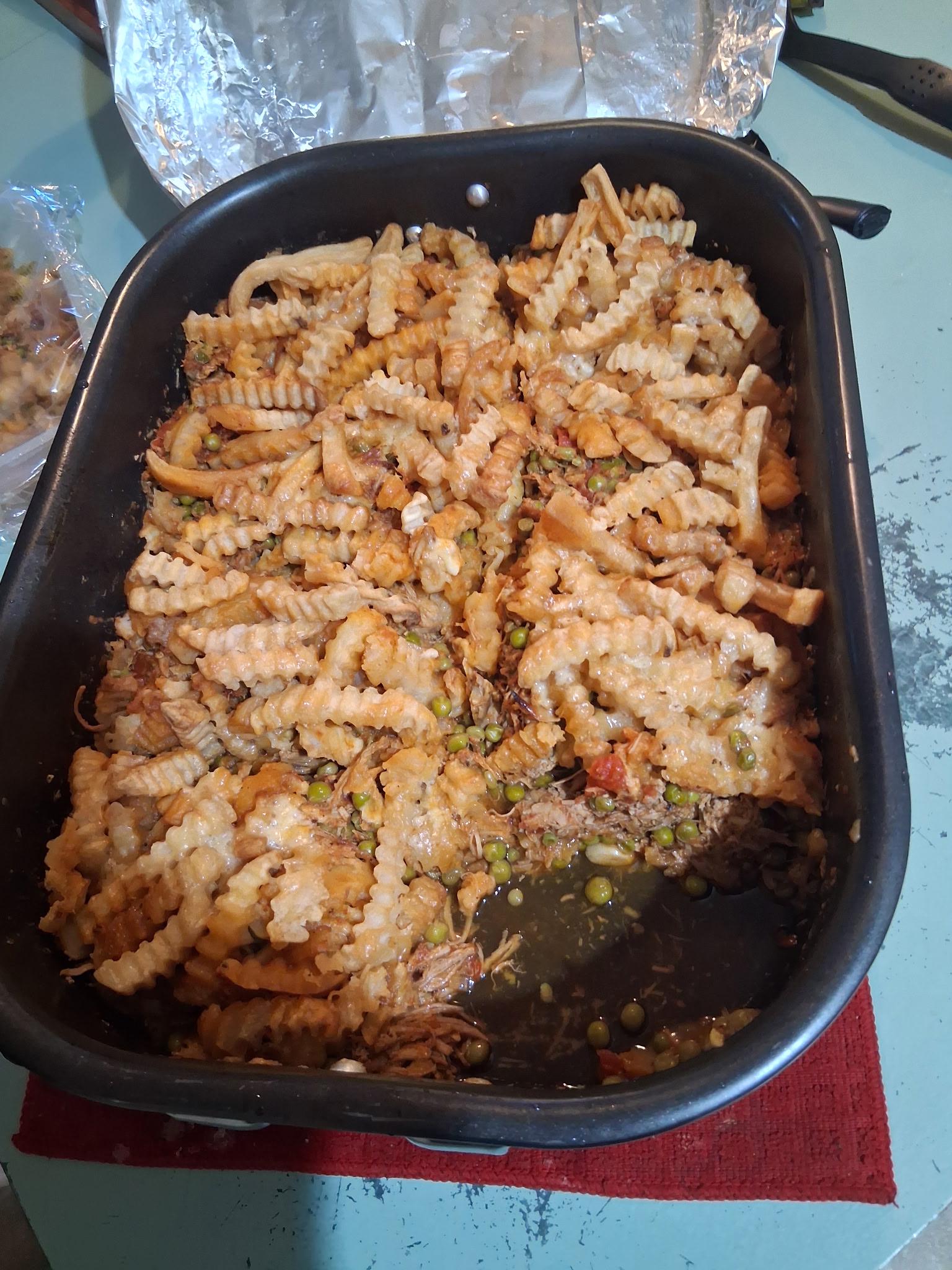A black roasting pan filled with crinkle-cut fries mixed with peas, shredded meat, and vegetables, resting on a red cloth on a blue table. Some liquid is visible at the bottom of the pan.
