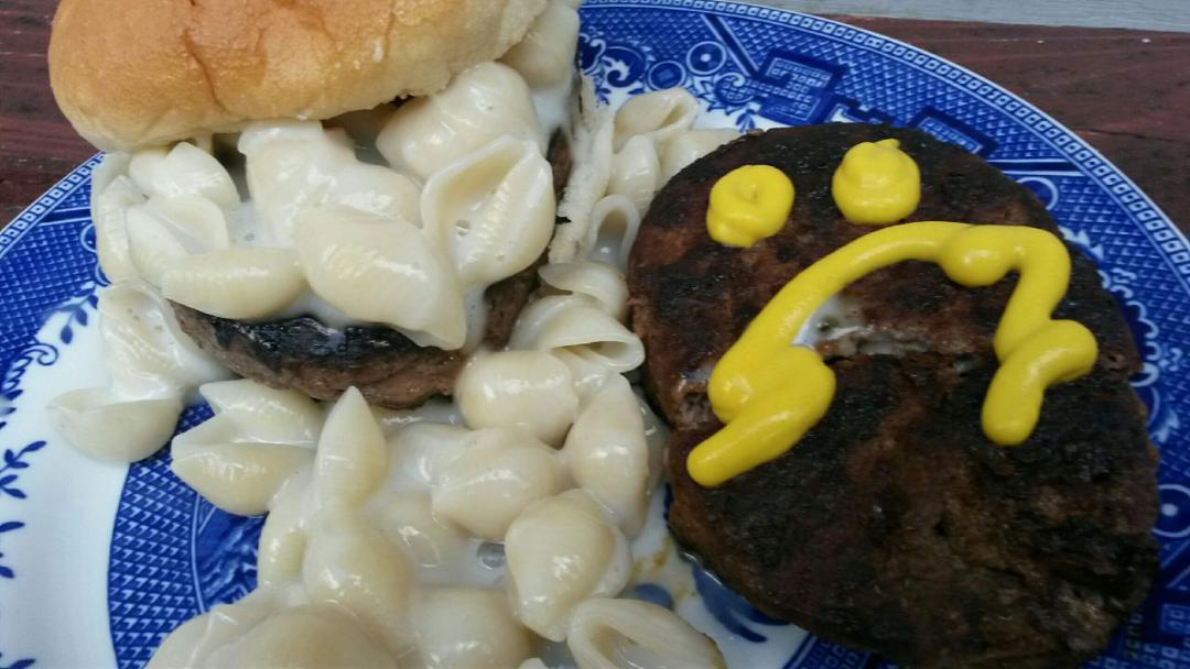 A burger with macaroni and cheese in a bun sits on a plate with extra macaroni and a beef patty topped with a sad face drawn in yellow mustard.