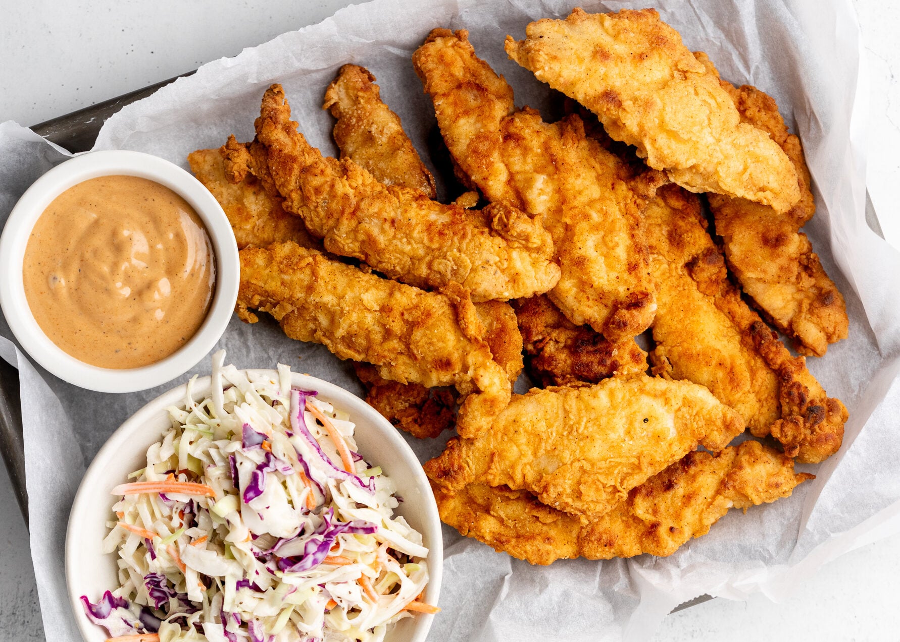 A tray of crispy fried chicken tenders on parchment paper, served with a bowl of creamy dipping sauce and a bowl of colorful coleslaw with cabbage and carrots—just like the crowd-pleasing favorites found at popular fried chicken chains.