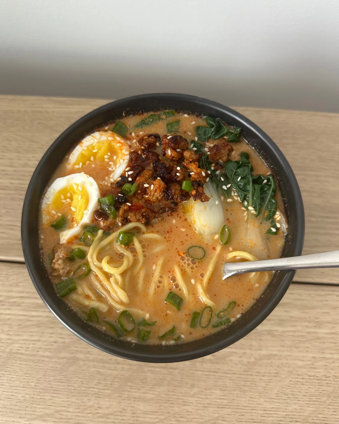 A bowl of ramen with half-boiled eggs, noodles, leafy greens, scallions, sesame seeds, and chili oil, served on a wooden surface with a spoon.