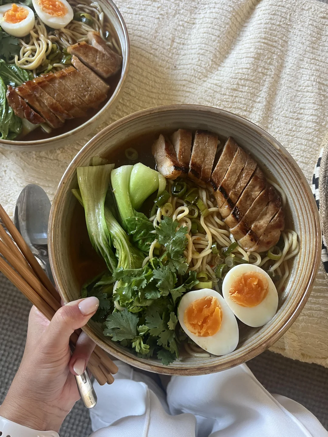A hand holds a bowl of ramen with noodles, bok choy, cilantro, sliced pork, green onions, and two halved soft-boiled eggs. Another similar bowl is partially visible in the background.