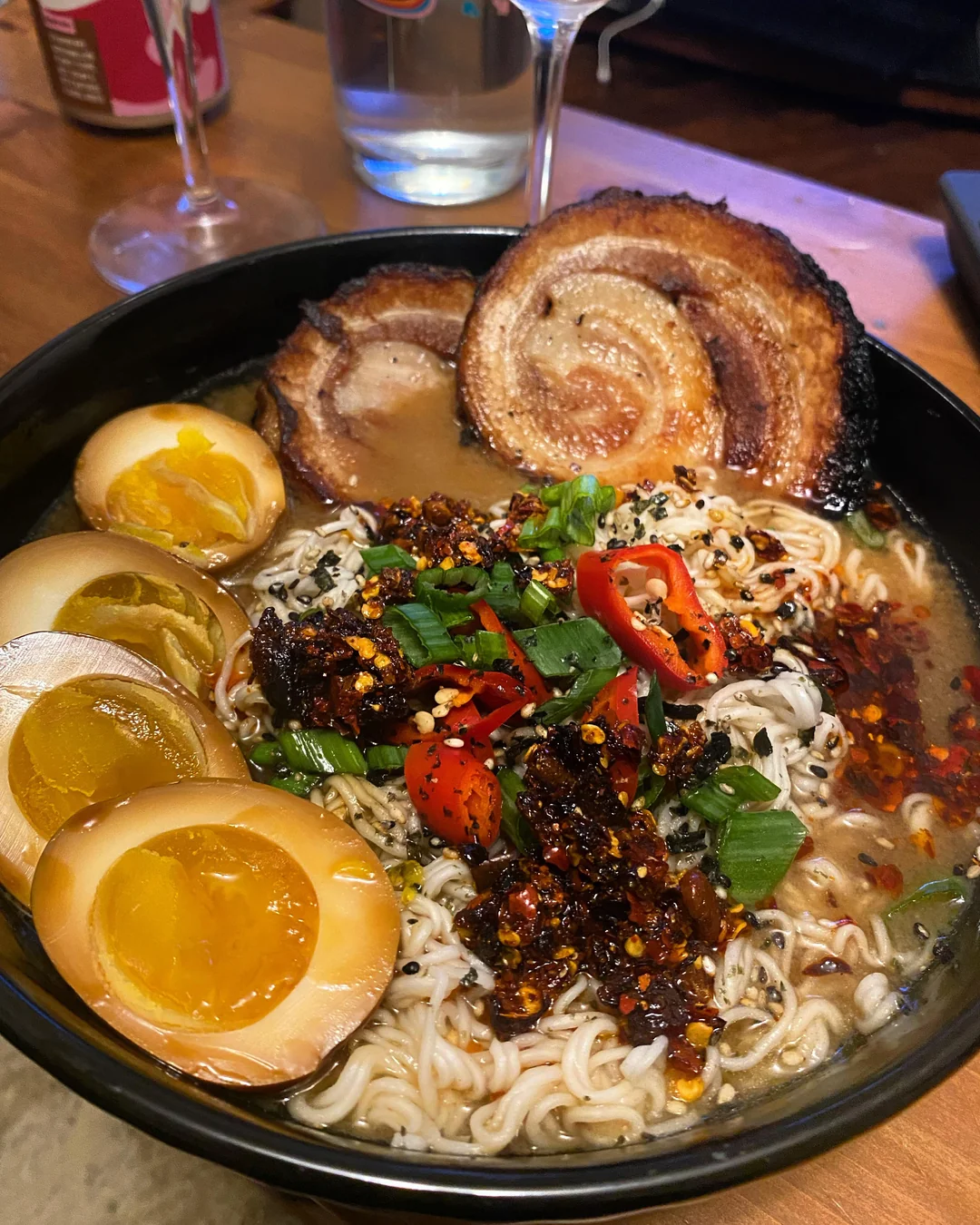 A bowl of ramen topped with two slices of pork belly, three halved marinated eggs, chopped green onions, red chilies, sesame seeds, and chili oil in rich broth, on a wooden table.