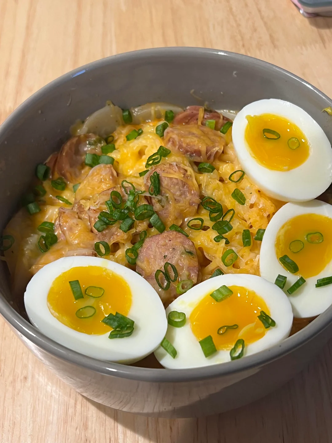 A bowl of noodles topped with sliced sausage, shredded cheese, chopped green onions, and three halved soft-boiled eggs with bright yellow yolks.