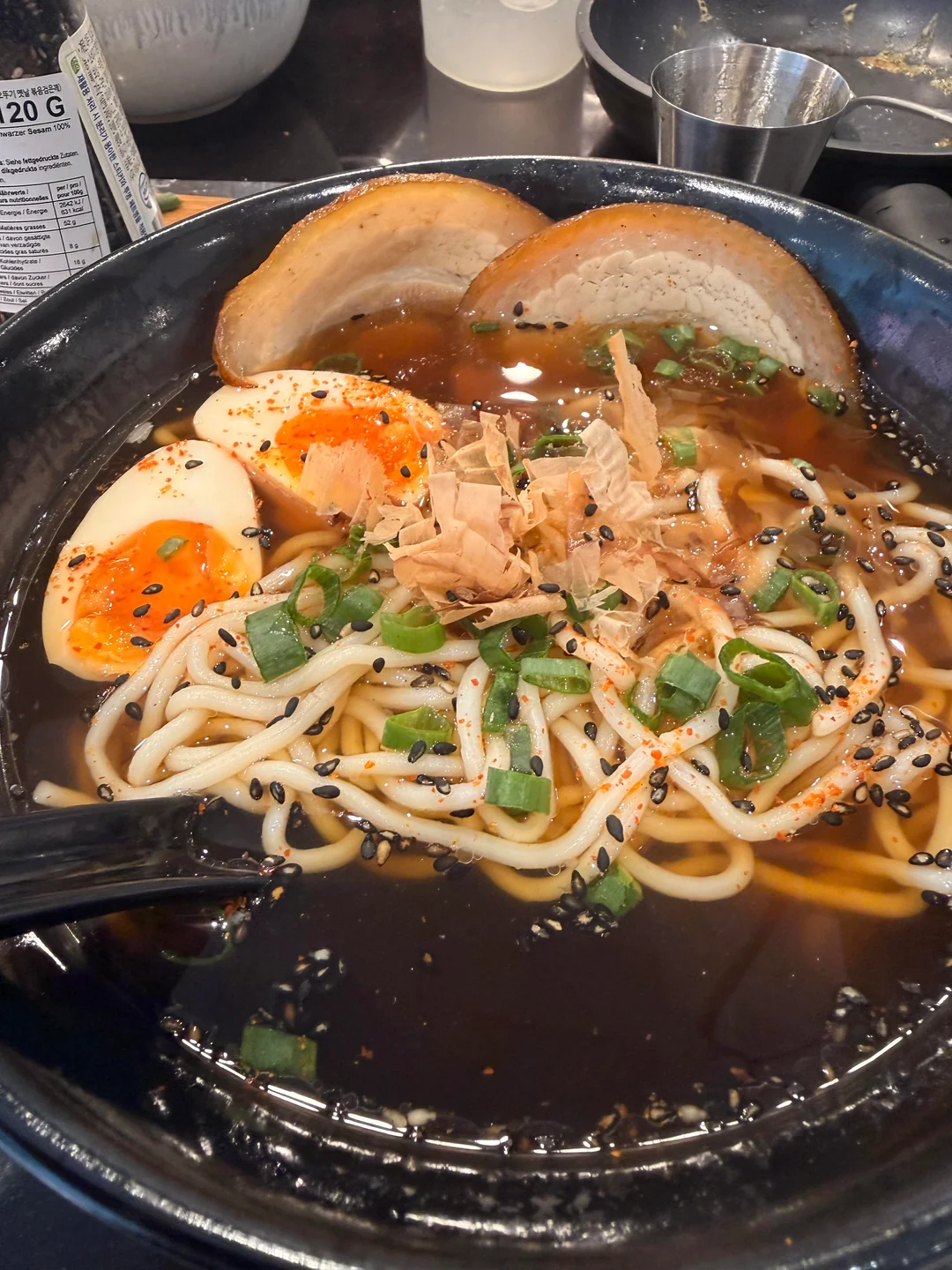 Bowl of ramen with sliced pork, soft-boiled eggs, green onions, bonito flakes, sesame seeds, and thick noodles in broth. A spoon rests on the bowl’s edge.