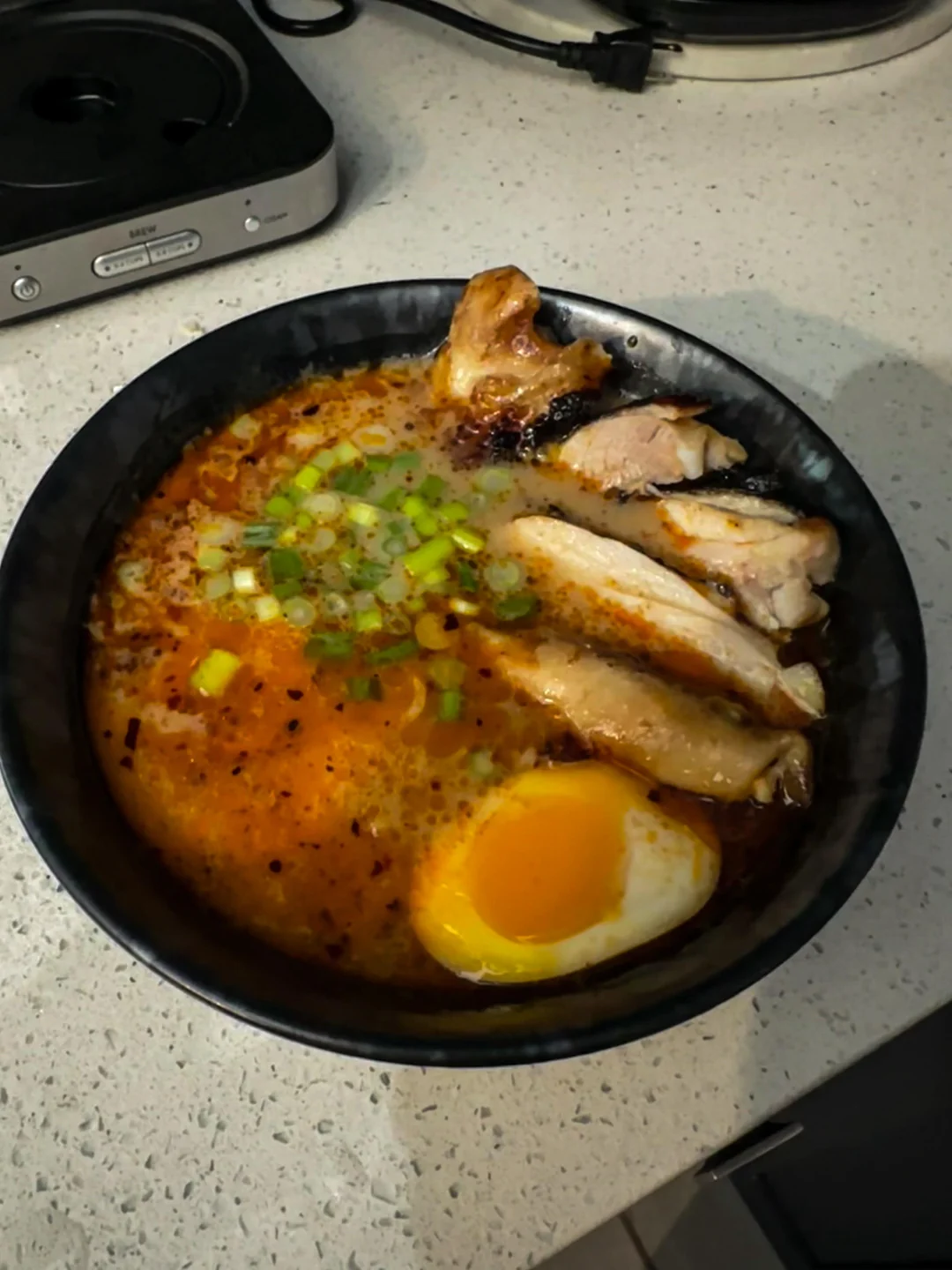 A bowl of ramen with sliced chicken, a soft-boiled egg, green onions, and spicy broth sits on a countertop.