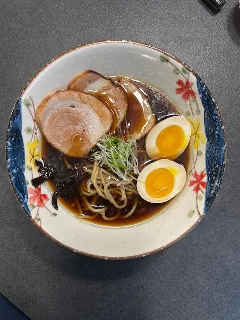 A bowl of ramen with sliced pork, soft-boiled egg halves, noodles, seaweed, green onions, and dark broth, served in a decorative bowl on a gray surface.
