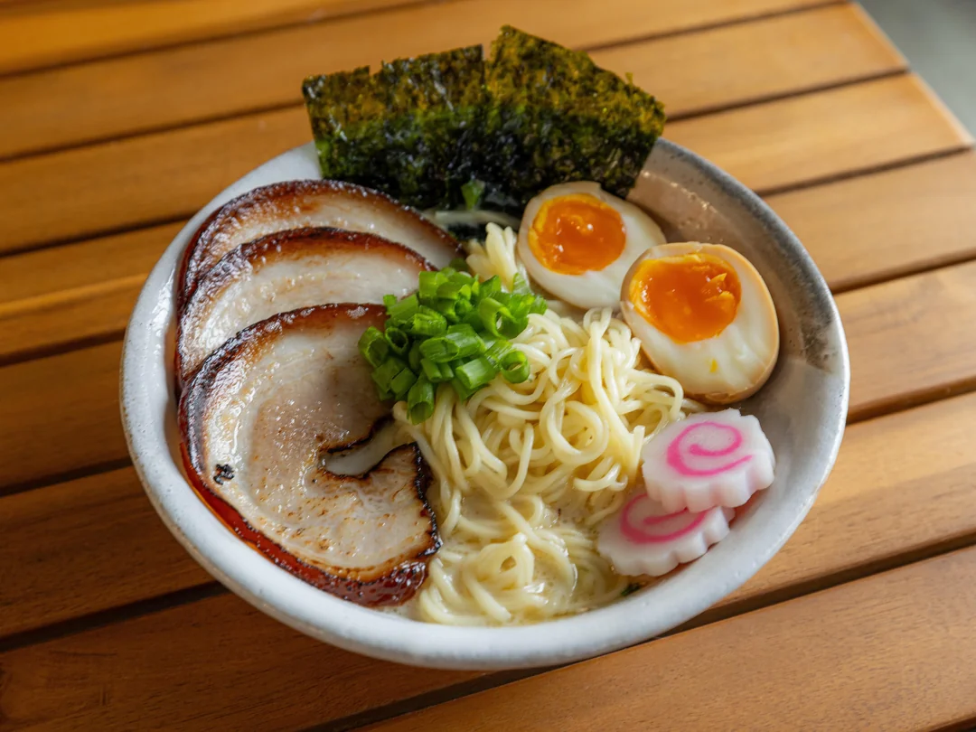 A bowl of ramen with sliced pork, halved soft-boiled eggs, narutomaki, green onions, seaweed, and noodles in broth sits on a wooden table.