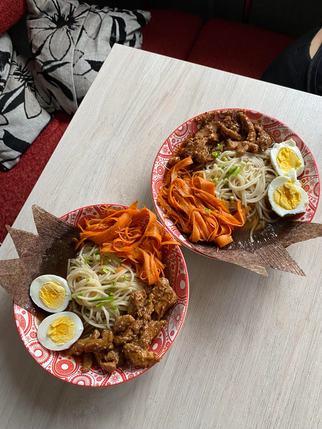 Two red patterned bowls on a light table, each filled with noodles, a halved boiled egg, shredded carrots, meat pieces, green onions, and brown crispy crackers, set near black-and-white floral pillows.