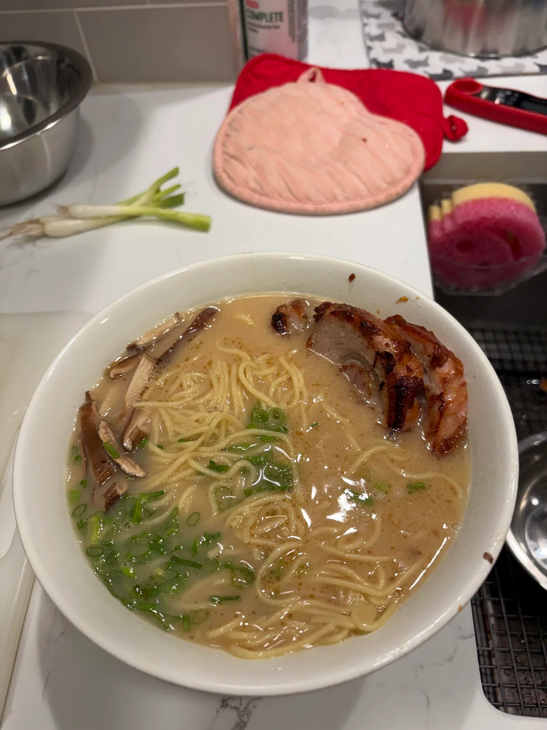 A bowl of ramen with sliced pork, green onions, and noodles in broth sits on a kitchen counter near a metal bowl, scallions, pink oven mitts, and a sink with a sponge.