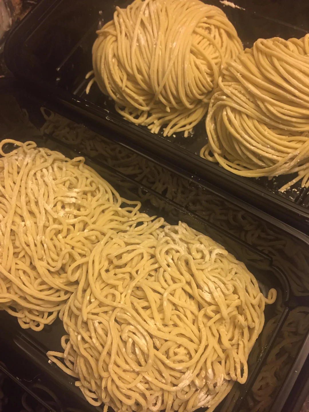Two portions of uncooked, hand-formed pasta noodles are placed in a black plastic container, with two neat nests on top and two looser piles on the bottom.