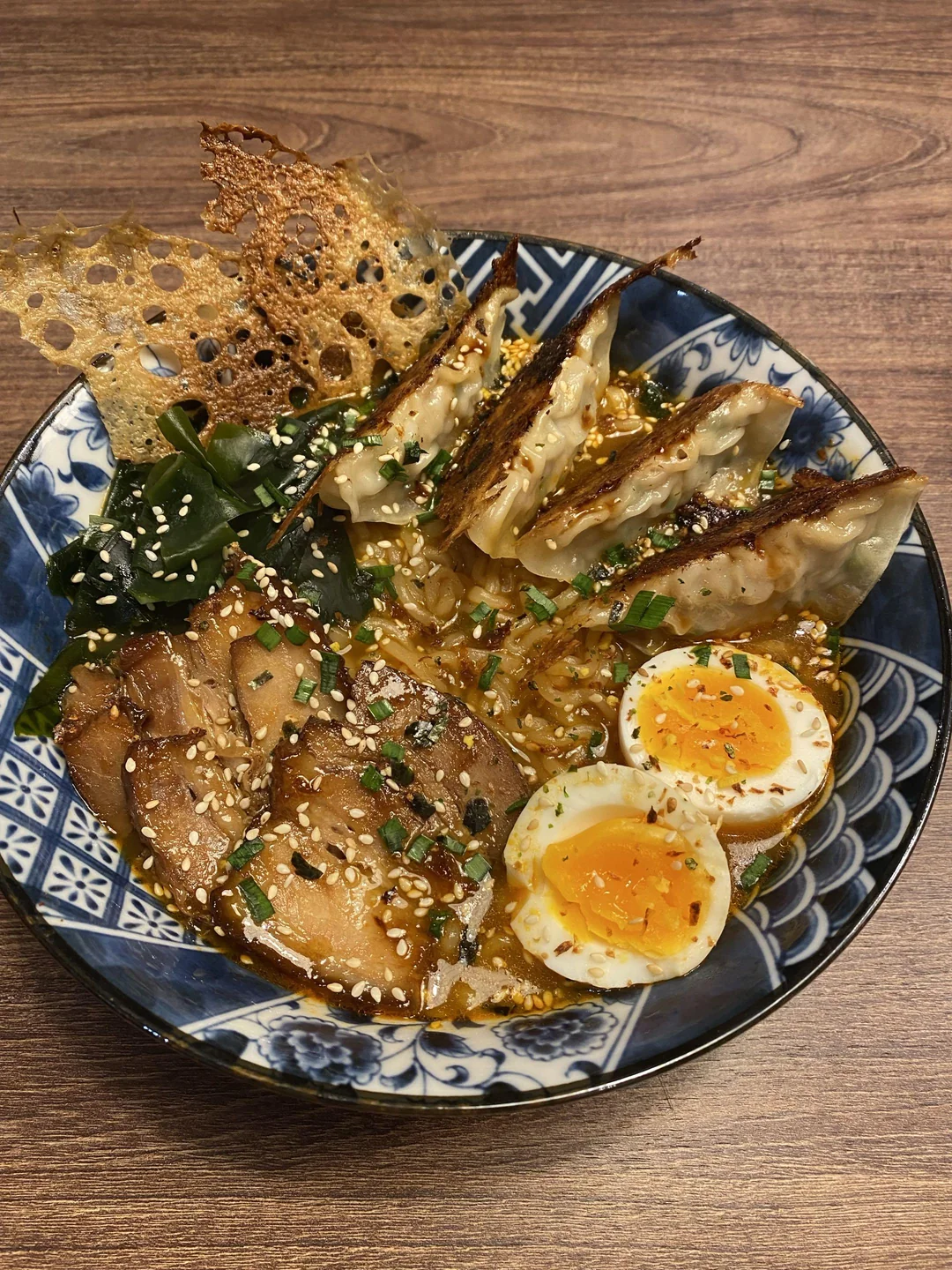 A bowl of ramen with sliced pork, soft-boiled egg halves, dumplings, seaweed, green onions, sesame seeds, and crispy garnish on top, served in a decorative blue bowl on a wooden table.