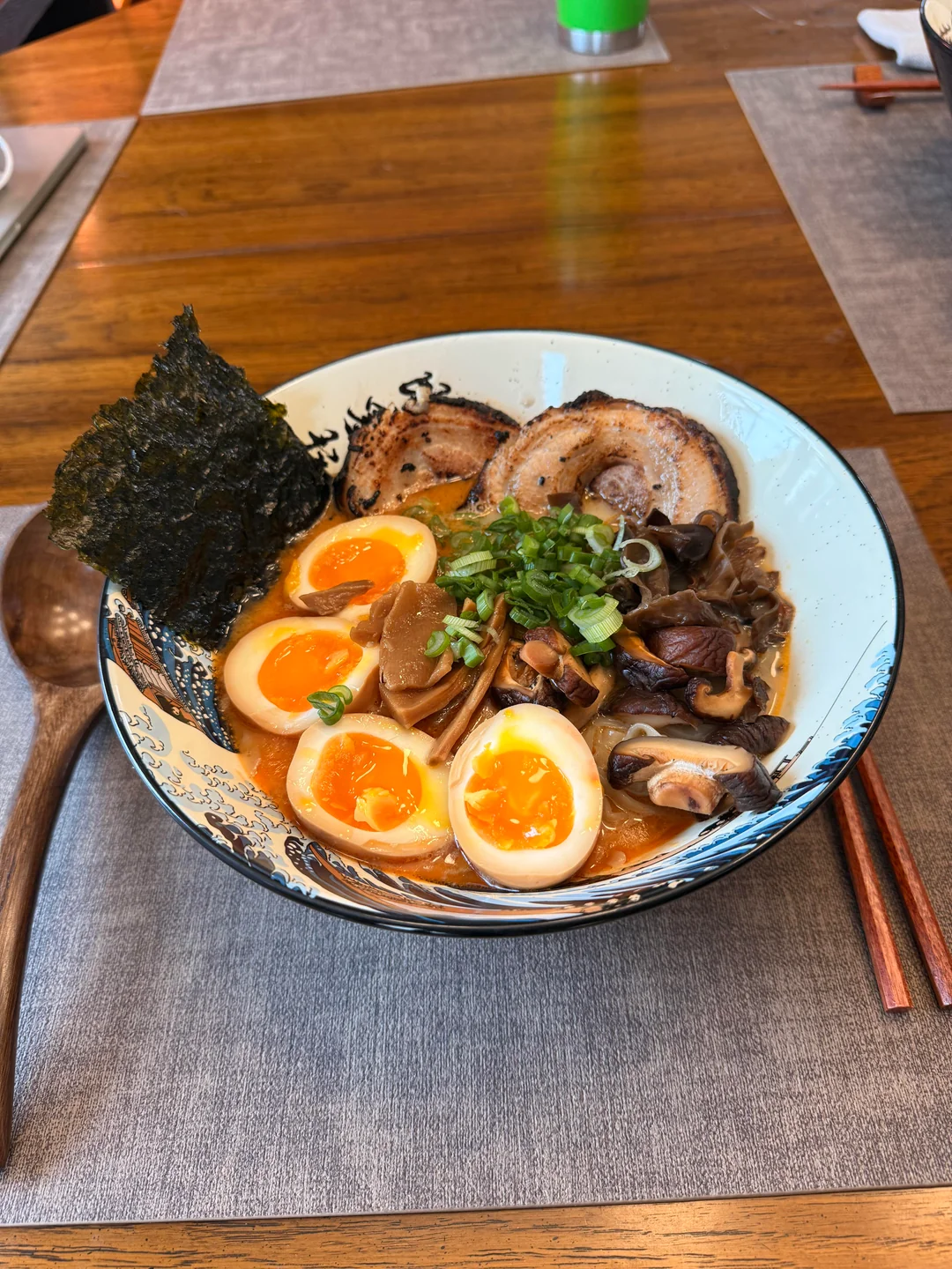 A bowl of ramen with sliced pork, half-boiled eggs, green onions, mushrooms, bamboo shoots, and a sheet of seaweed, placed on a table with chopsticks and a wooden spoon.