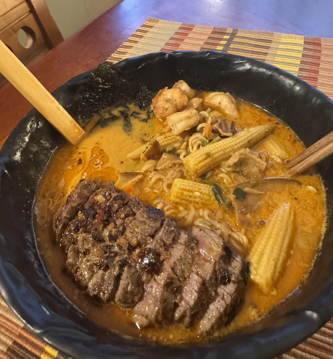 A bowl of spicy ramen with sliced beef, baby corn, mushrooms, and seafood in a rich broth, served with wooden chopsticks on a woven placemat.
