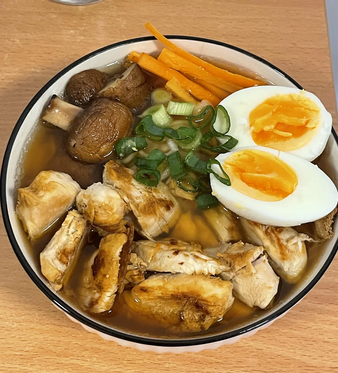 A bowl of ramen topped with sliced grilled chicken, mushrooms, carrot sticks, green onions, and a halved soft-boiled egg on a wooden table.