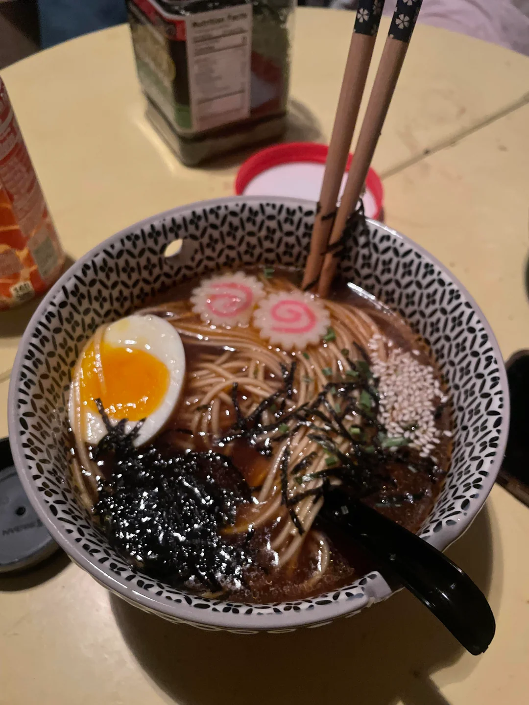 A bowl of ramen with broth, noodles, seaweed, sesame seeds, narutomaki slices, half a soft-boiled egg, and chopsticks resting on top. A spoon is in the bowl, and a can and container are in the background.