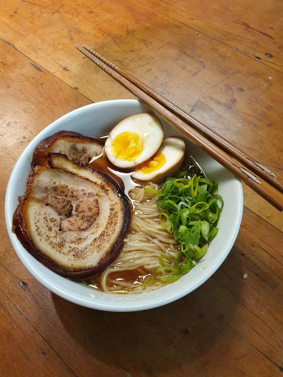 A bowl of ramen with sliced pork, soft-boiled egg, green onions, and noodles in broth. Chopsticks rest on the bowl, which sits on a wooden table.